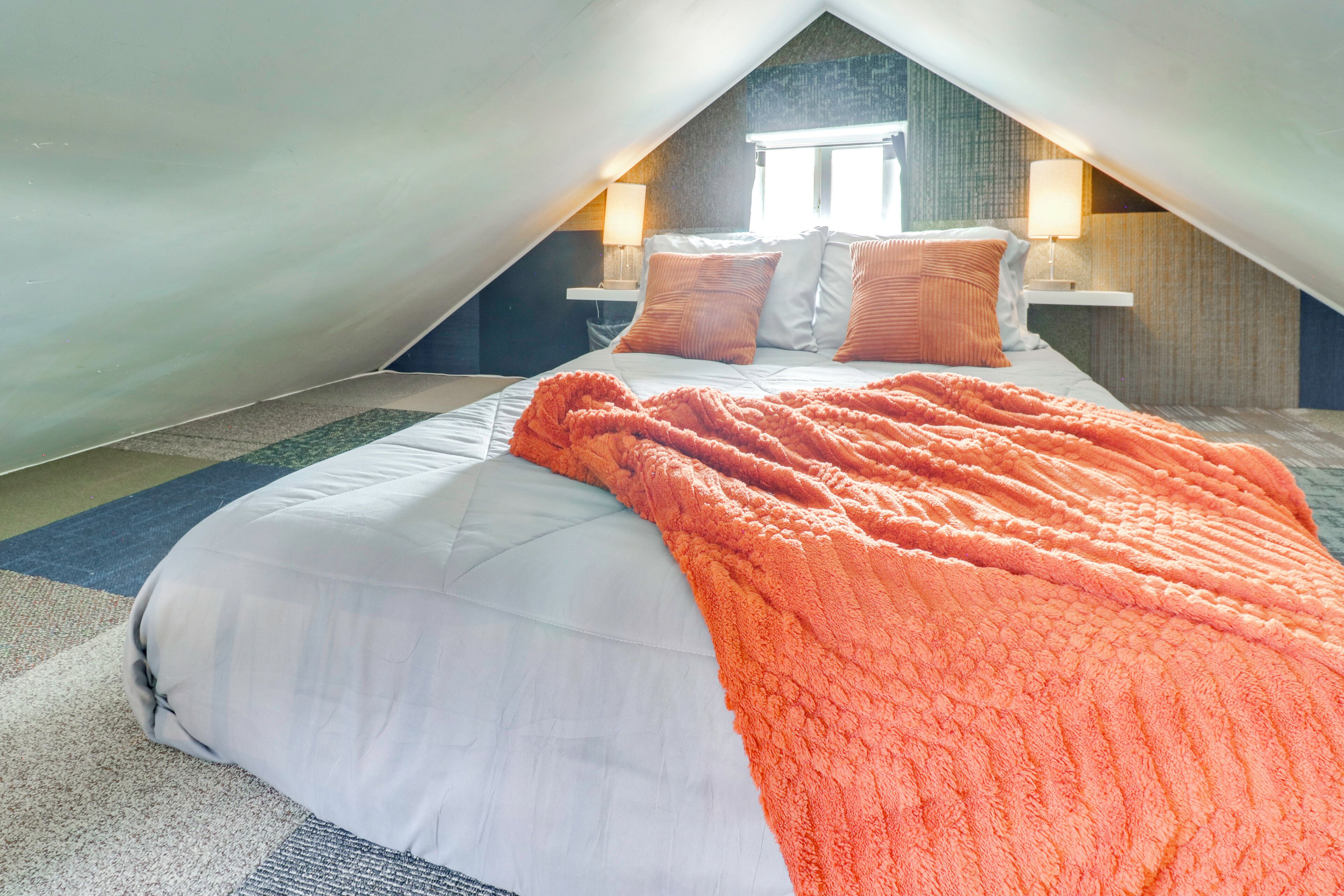 Een slaapkamer met een schuine plafond, een bed met wit beddengoed en een oranje deken, twee bedlampjes en een raam.