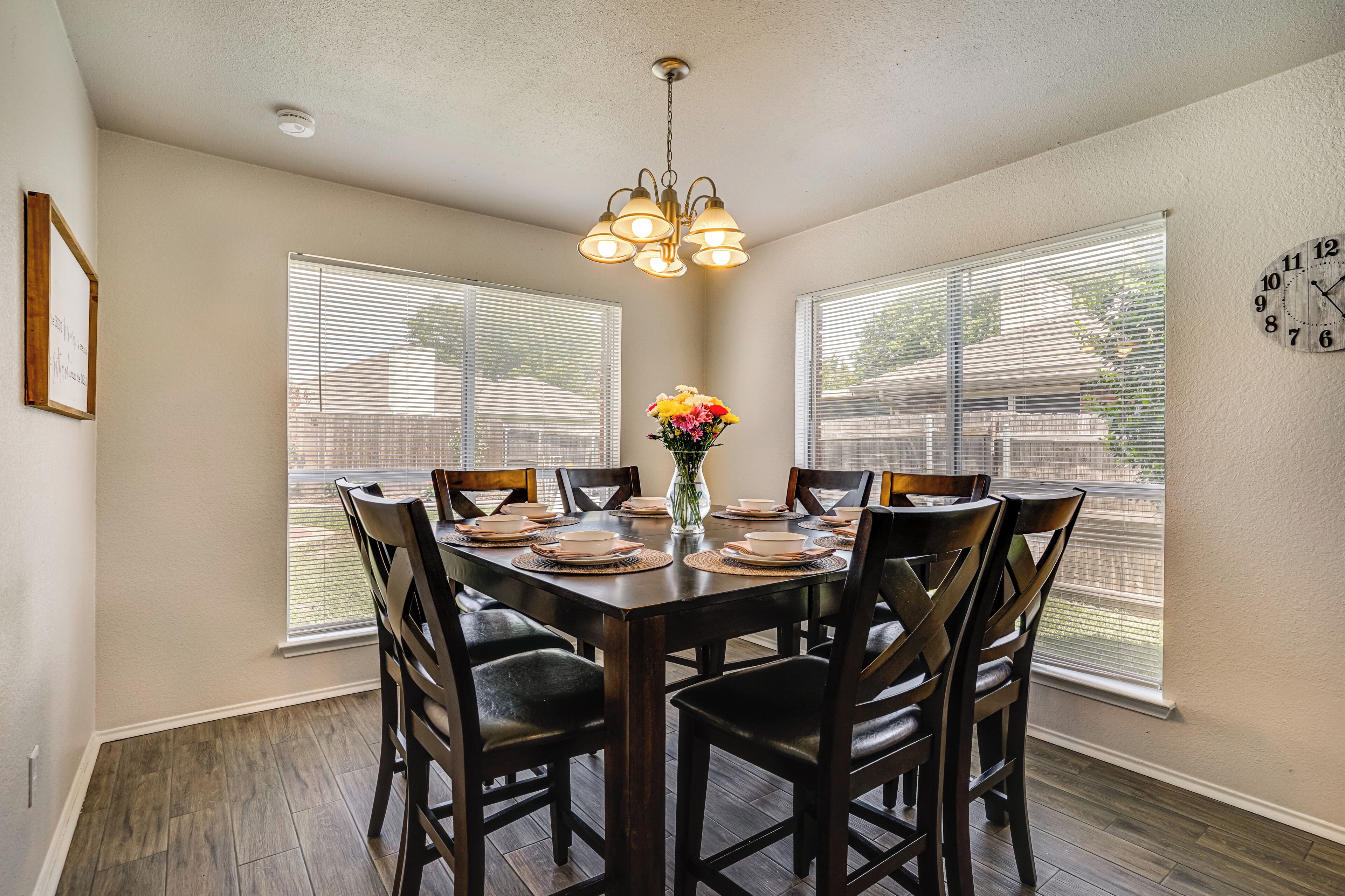 A dining room with a table set for six, surrounded by chairs, a chandelier, and a clock on the wall.