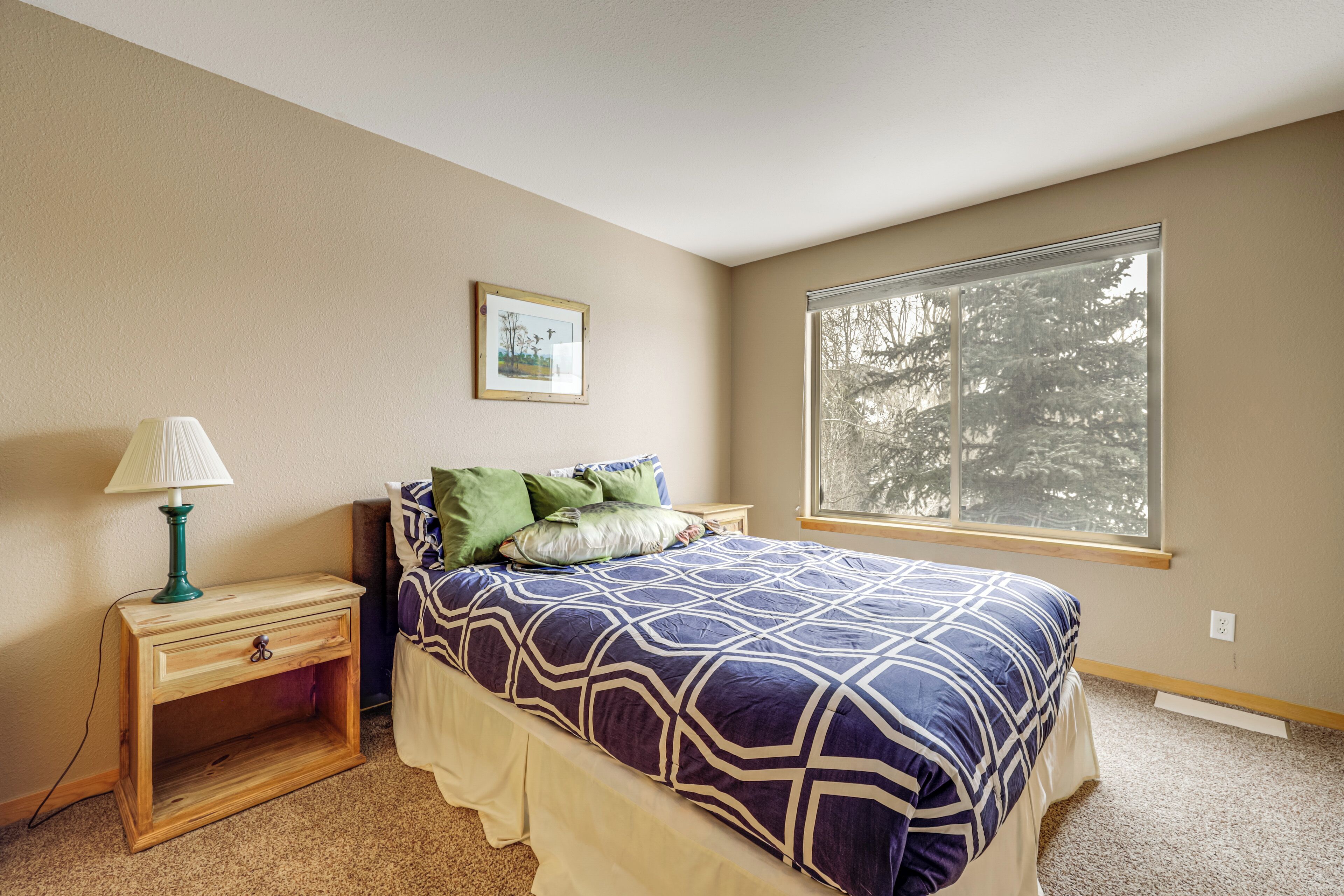 A bedroom with a bed, a nightstand, a lamp, a window with a view of trees, and a framed picture on the wall.