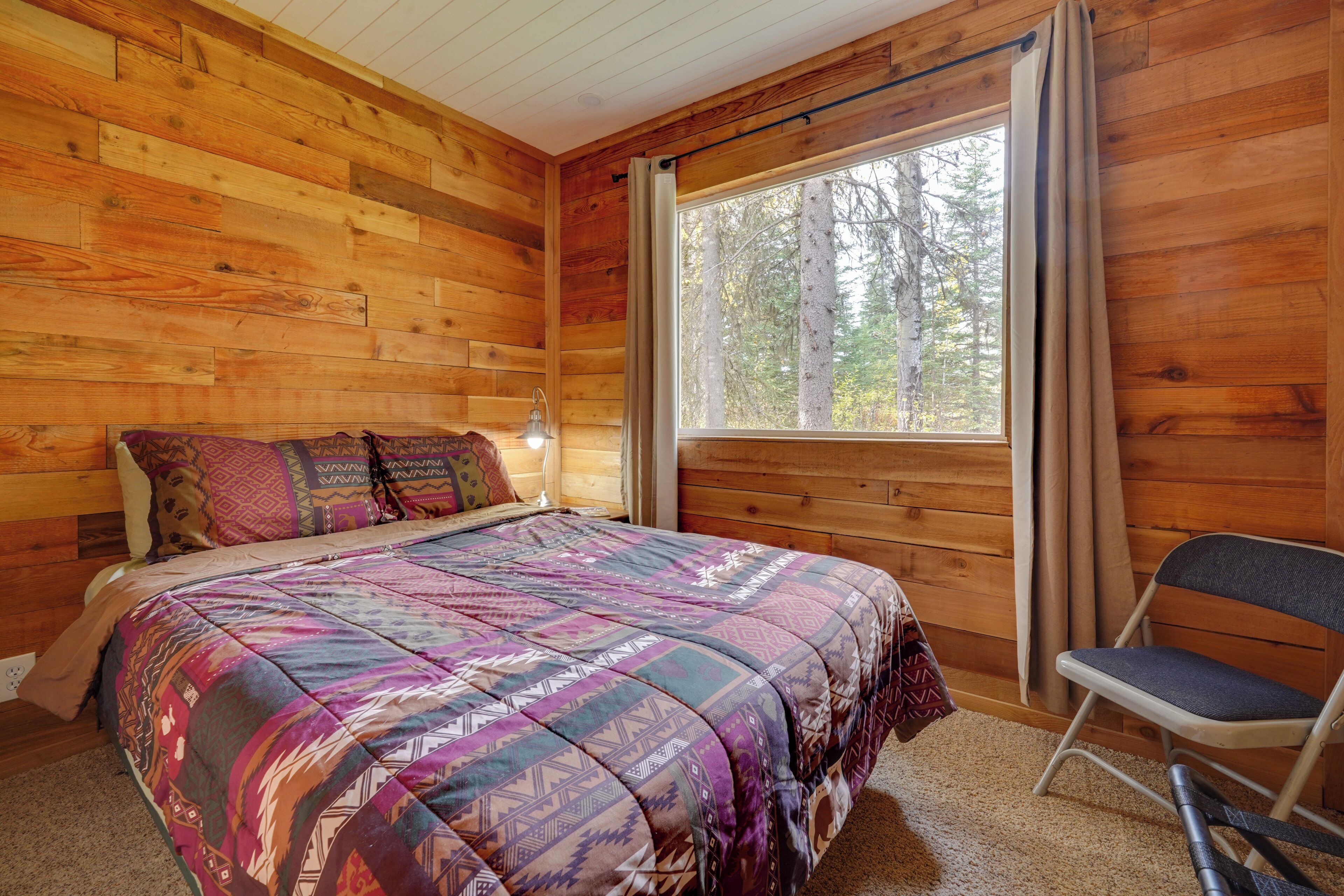 A wooden cabin room with a bed, a chair, and a window with curtains.