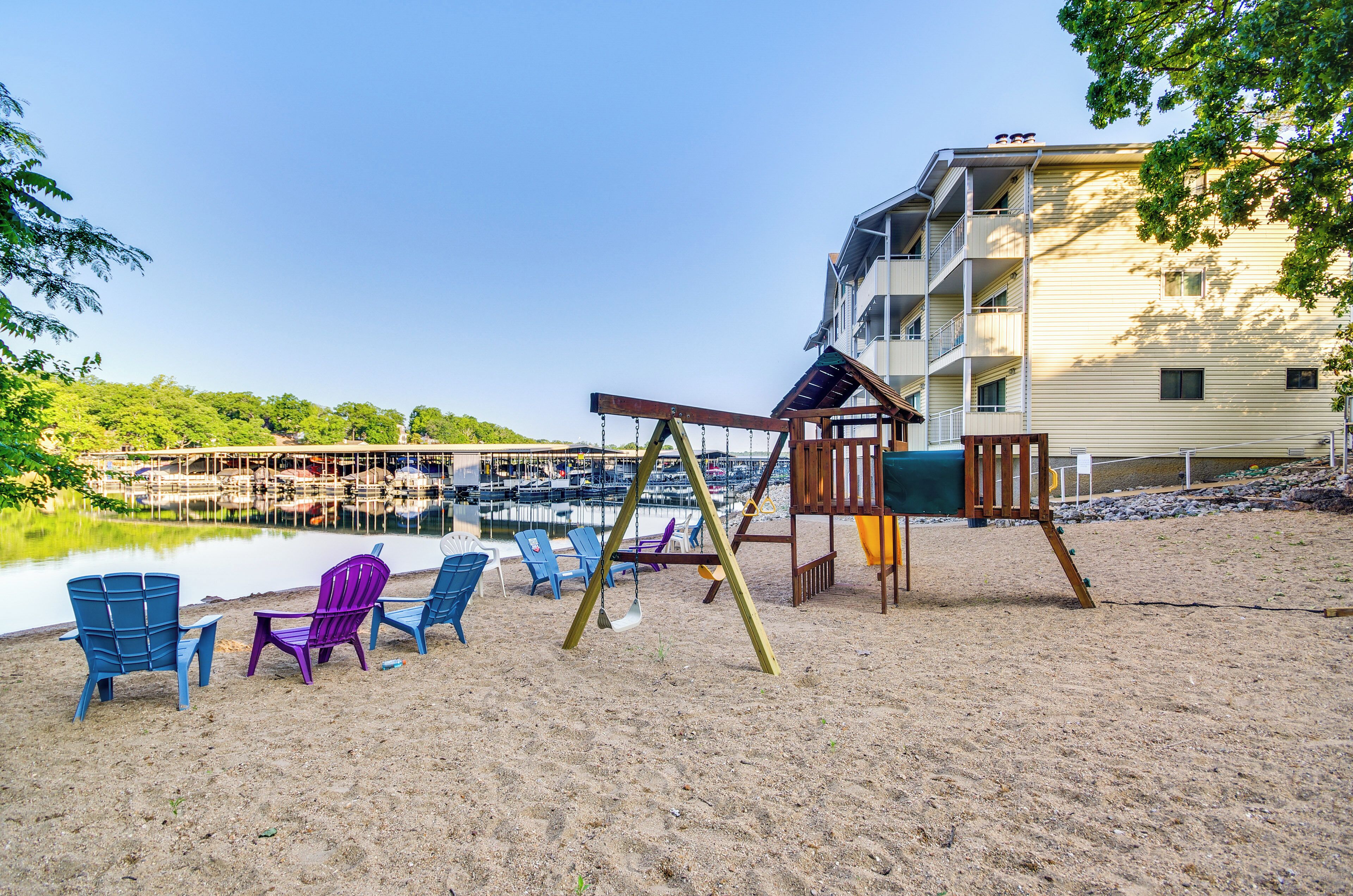 Een zandstrand met een schommel en kleurrijke Adirondack-stoelen. Op de achtergrond een gebouw met meerdere balkons en een steiger met boten.