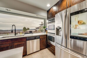 A modern kitchen with stainless steel appliances, wooden cabinets, and a large window overlooking a residential area. - Puget Sound Views! Serene Federal Way Retreat (Federal Way)
