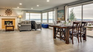 Amplia sala de estar con chimenea, comedor con mesa y sillas de madera, ventanas amplias con vista al agua y un rincón de descanso acogedor con sofá y sillón.