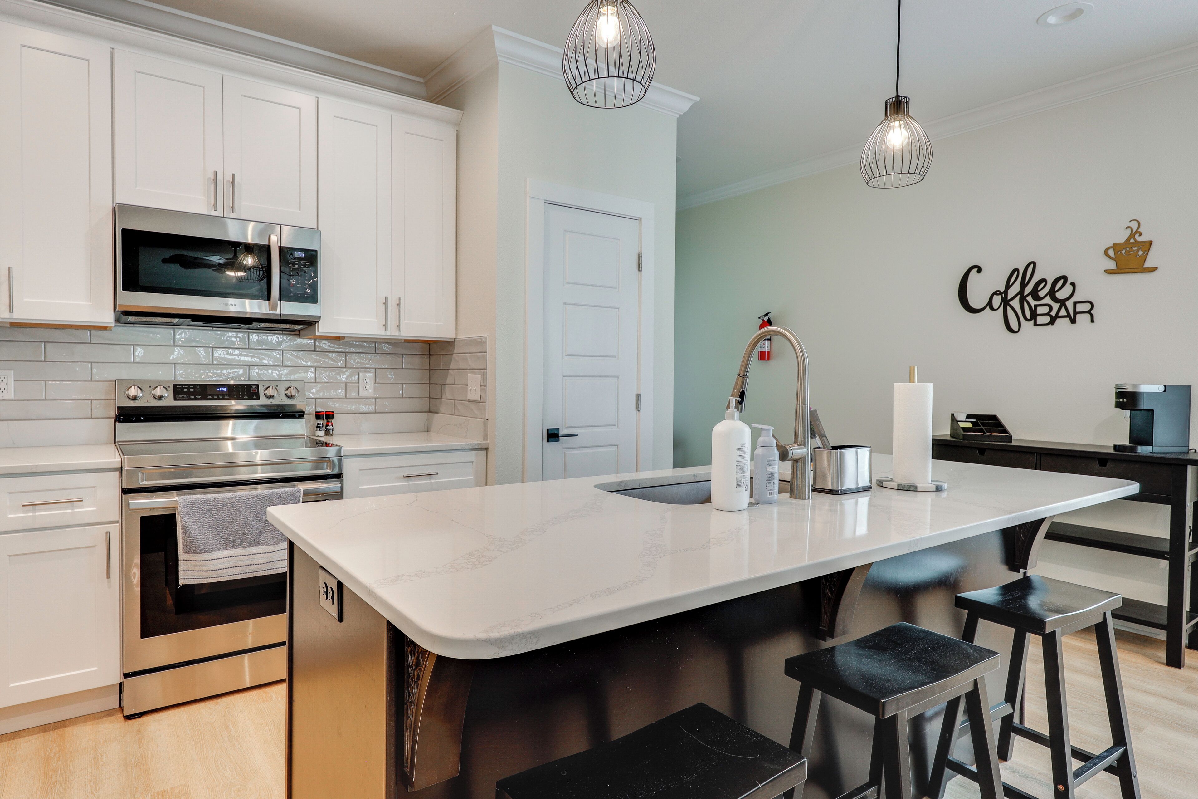 A modern kitchen with white cabinets, stainless steel appliances, a large island with stools, and a 'Coffee Bar' sign.