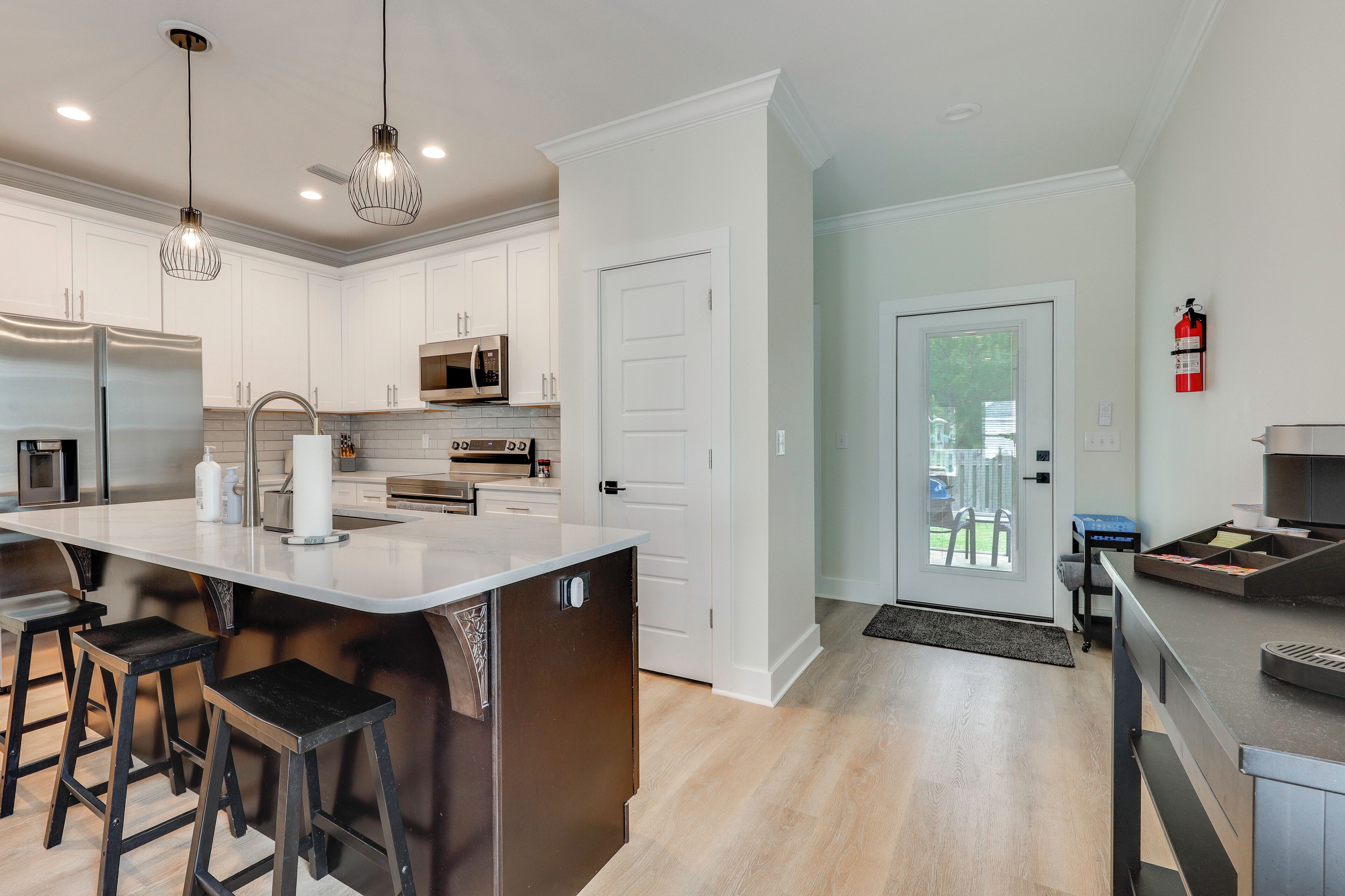 A modern kitchen with white cabinets, stainless steel appliances, a large island with stools, pendant lighting, and a fire extinguisher on the wall.