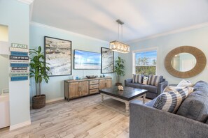 A living room with a sofa, coffee table, and a large mirror. There are framed pictures on the wall and a sign with beach-related phrases. - Introducing Shells N Sea Beach Villa at Lost Key Golf & Beach Club! 4BR 4BA (Pensacola)