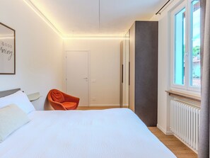 Quarto com cama, cadeira, guarda-roupa, radiador e janela com vista.