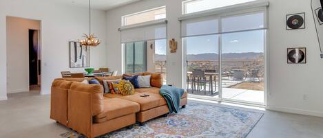 A spacious living room with a large sectional sofa, a chandelier, and a view of the outdoors through large sliding glass doors.