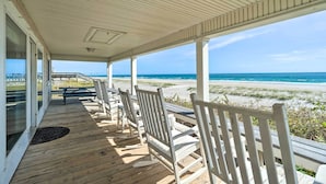 Une terrasse couverte avec un plancher en bois et des rampes blanches, offrant une vue dégagée sur la plage et l’océan.