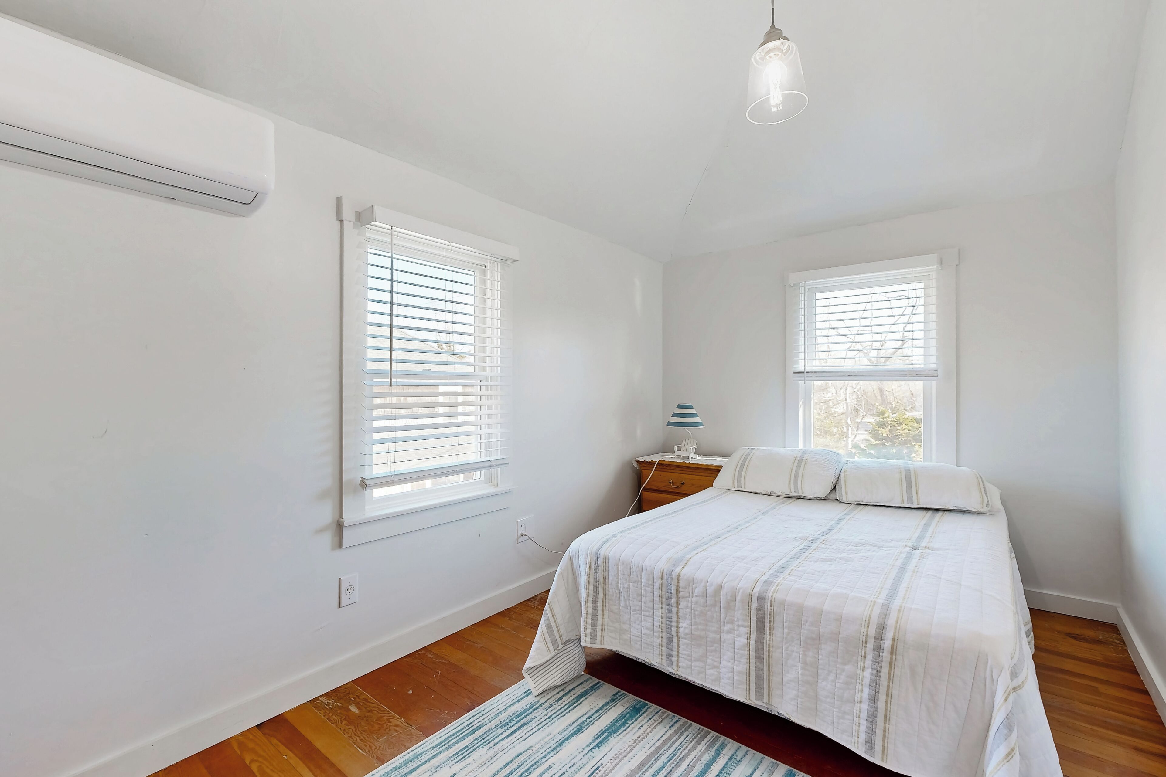A bedroom with a bed, two windows with blinds, a bedside table with a lamp, and a hanging light fixture.