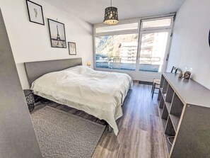 Ein Schlafzimmer mit einem Bett, einem Schreibtisch, einem Fenster mit Blick auf Gebäude und gerahmten Bildern an der Wand.