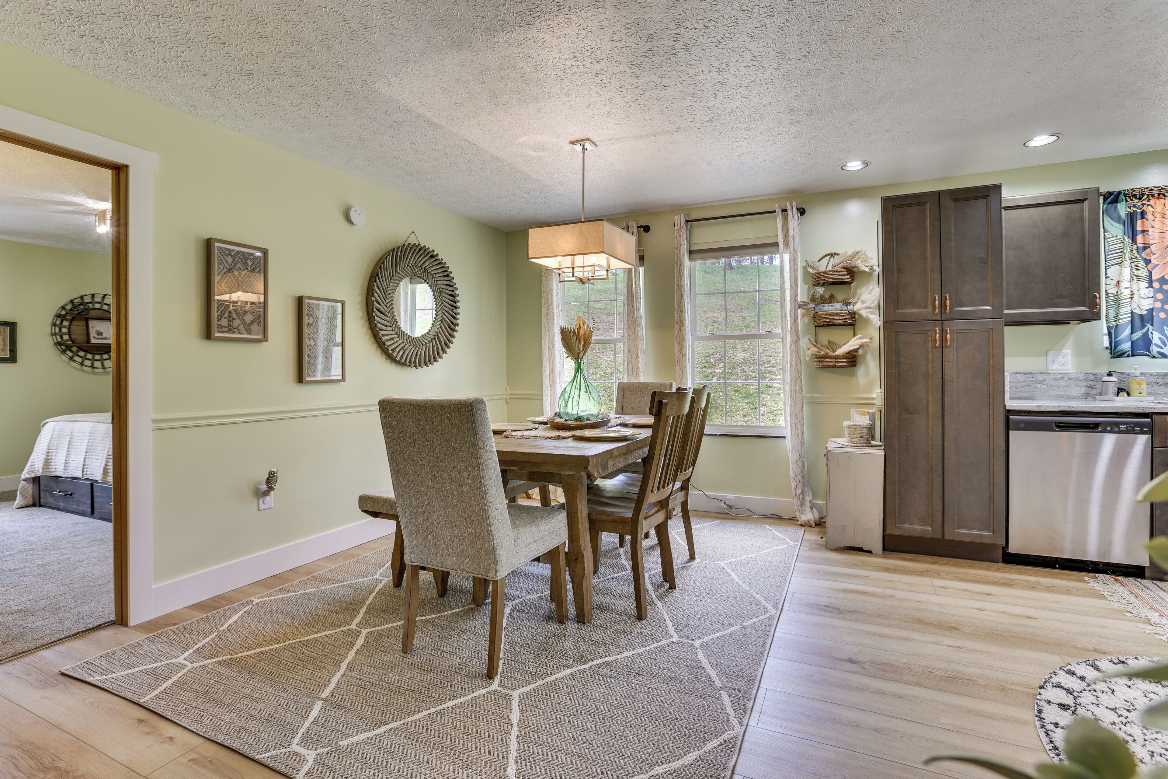 A dining area with a wooden table and chairs, a hanging light fixture, and a kitchen with a dishwasher and cabinets.