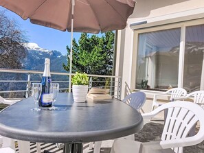 Uma mesa redonda com uma garrafa, um copo e um livro, rodeada por cadeiras brancas, com vista para a montanha ao fundo.