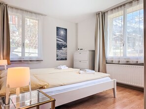 Quarto com cama, mesas de cabeceira, cômoda e janela com cortinas.