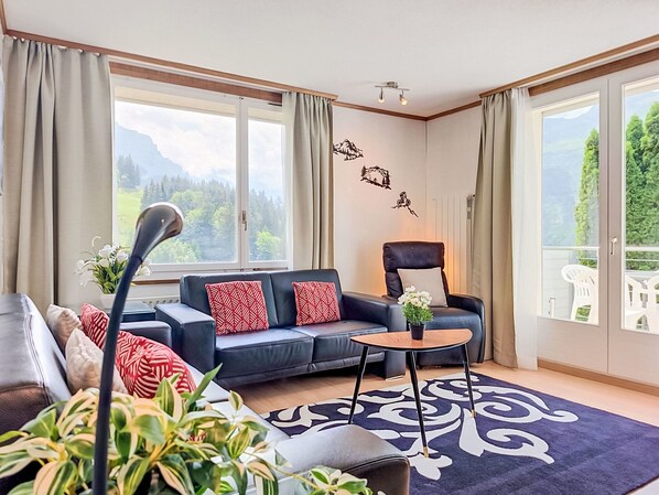 Uma sala de estar moderna com sofá, poltrona e uma mesa pequena. Há grandes janelas com cortinas, vista para área verde e montanhas.