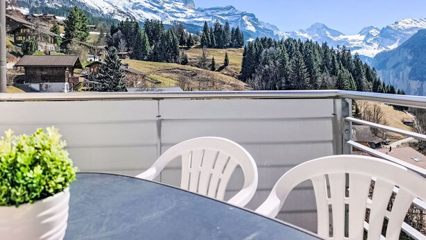 Un balcón con mesa y sillas, con vistas a un paisaje montañoso de picos nevados y bosques densos.