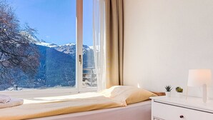Un dormitorio con cama, ventana con cortinas, mesita de noche con lámpara y una planta pequeña.