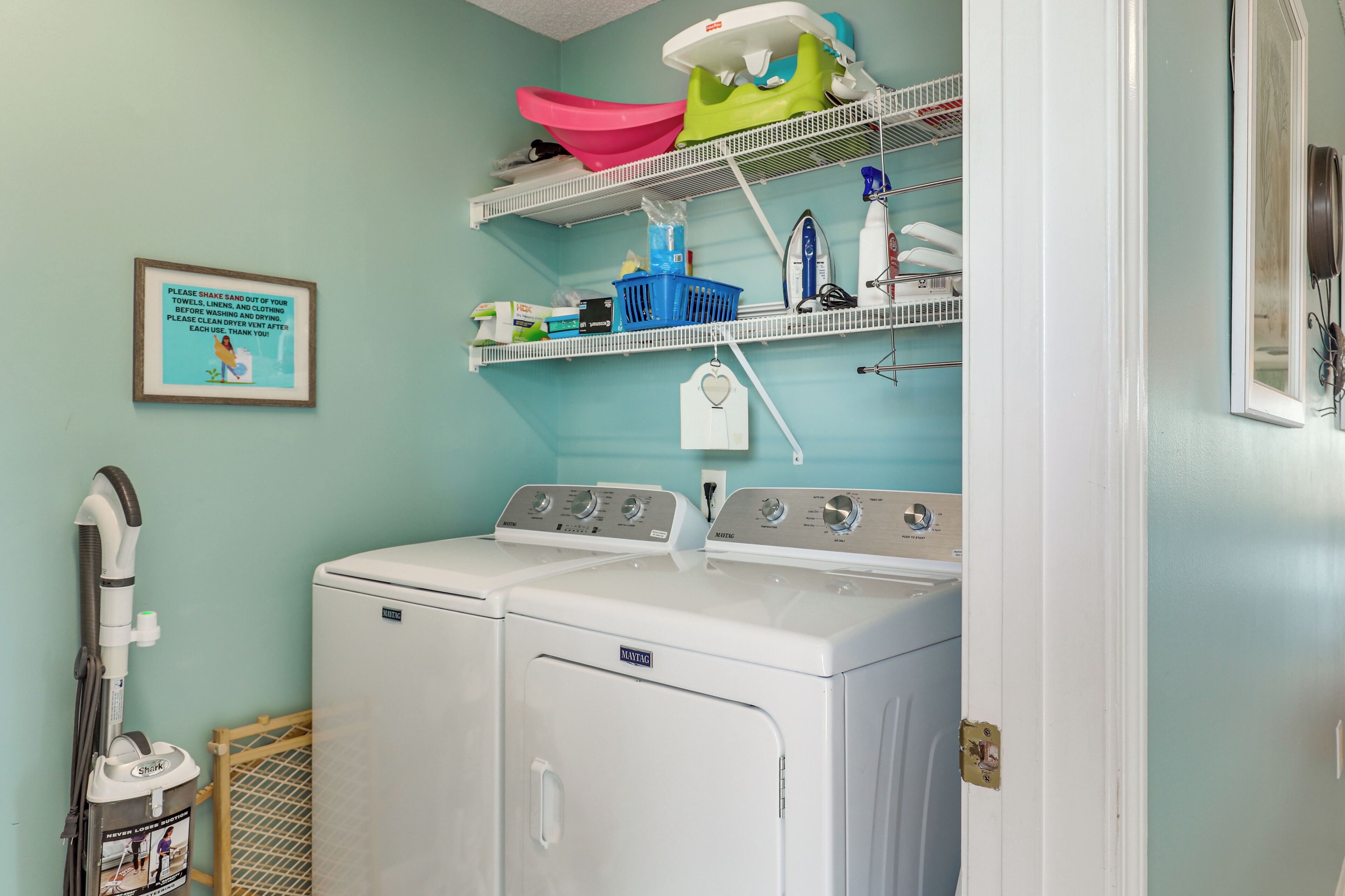 A laundry area with a front-loading washer and dryer, shelves with laundry supplies, and a framed safety message on the wall.