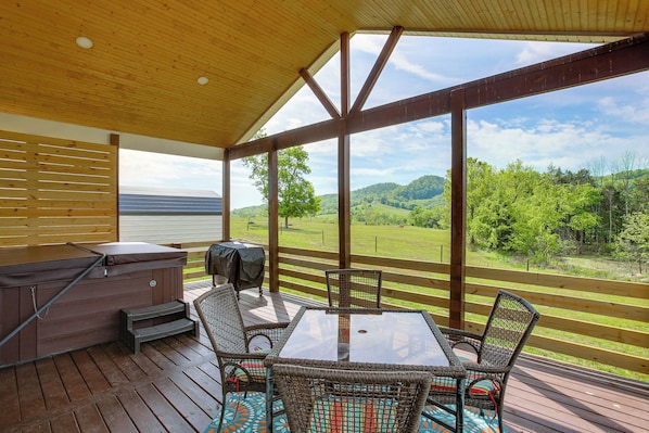 A covered outdoor deck with a table and chairs, a hot tub, and a view of a green landscape.