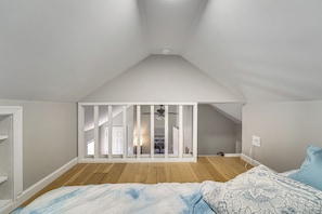 A bedroom with a sloped ceiling, wooden flooring, a bed with patterned bedding, and a view into an adjacent room through a white partition.