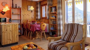 A cozy cabin interior with a dining area, wooden furniture, a large window with curtains, and a view of the mountains. - Beautiful apartment for 5 people with TV and balcony (Veysonnaz)