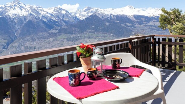 A balcony with a table set for two, a potted plant, and a view of snow-capped mountains. - Beautiful apartment for 5 people with TV and balcony (Veysonnaz)