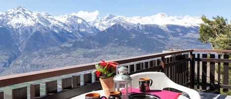 A balcony with a table set for two, a potted plant, and a view of snow-capped mountains.