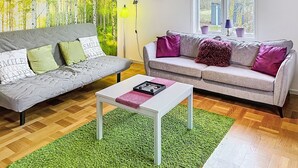 Un salon avec deux canapés, une table basse, un tapis vert, un tableau représentant des arbres et une fenêtre donnant sur un beau paysage.