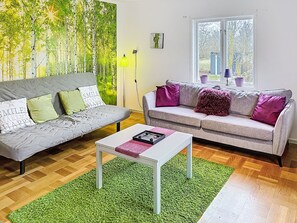 Een woonkamer met twee banken, een salontafel, een groen tapijt, een schilderij van bomen, en een raam met uitzicht.
