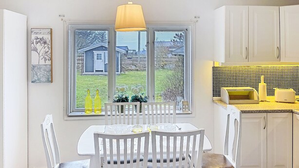 Une cuisine avec des meubles blancs, une table à manger avec des chaises, une fenêtre donnant sur une maison et un jardin, et un luminaire suspendu jaune.