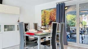 A modern dining area with a glass table, metal chairs, and a painting of trees. - La Maison des Palmiers by Interhome (Le Bouveret)