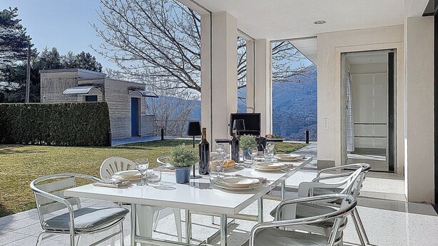 Un espacio de comedor al aire libre con una mesa lista para seis personas, rodeada de sillas con cojines, un televisor y una botella de vino.