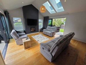 Ein modernes Wohnzimmer mit einem Flachbildfernseher, grauen Sofas, einem Kamin und großen Fenstern.