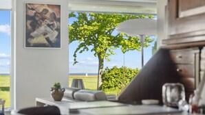 A modern living room with a view of the sea, a painting on the wall, and a dining area with a table and chairs.