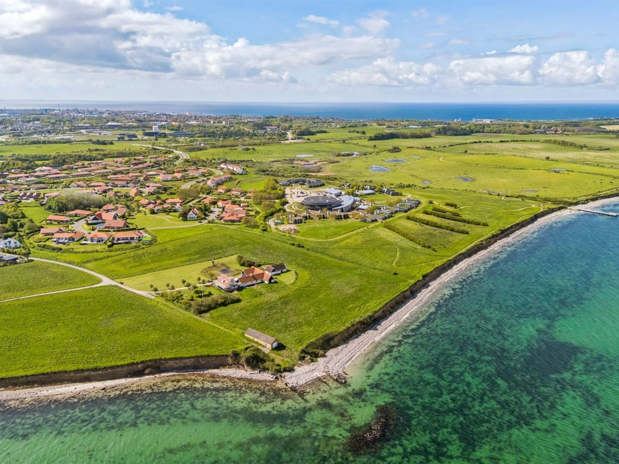 An aerial view of a coastal residential area with houses, green fields, and a clear coastline.