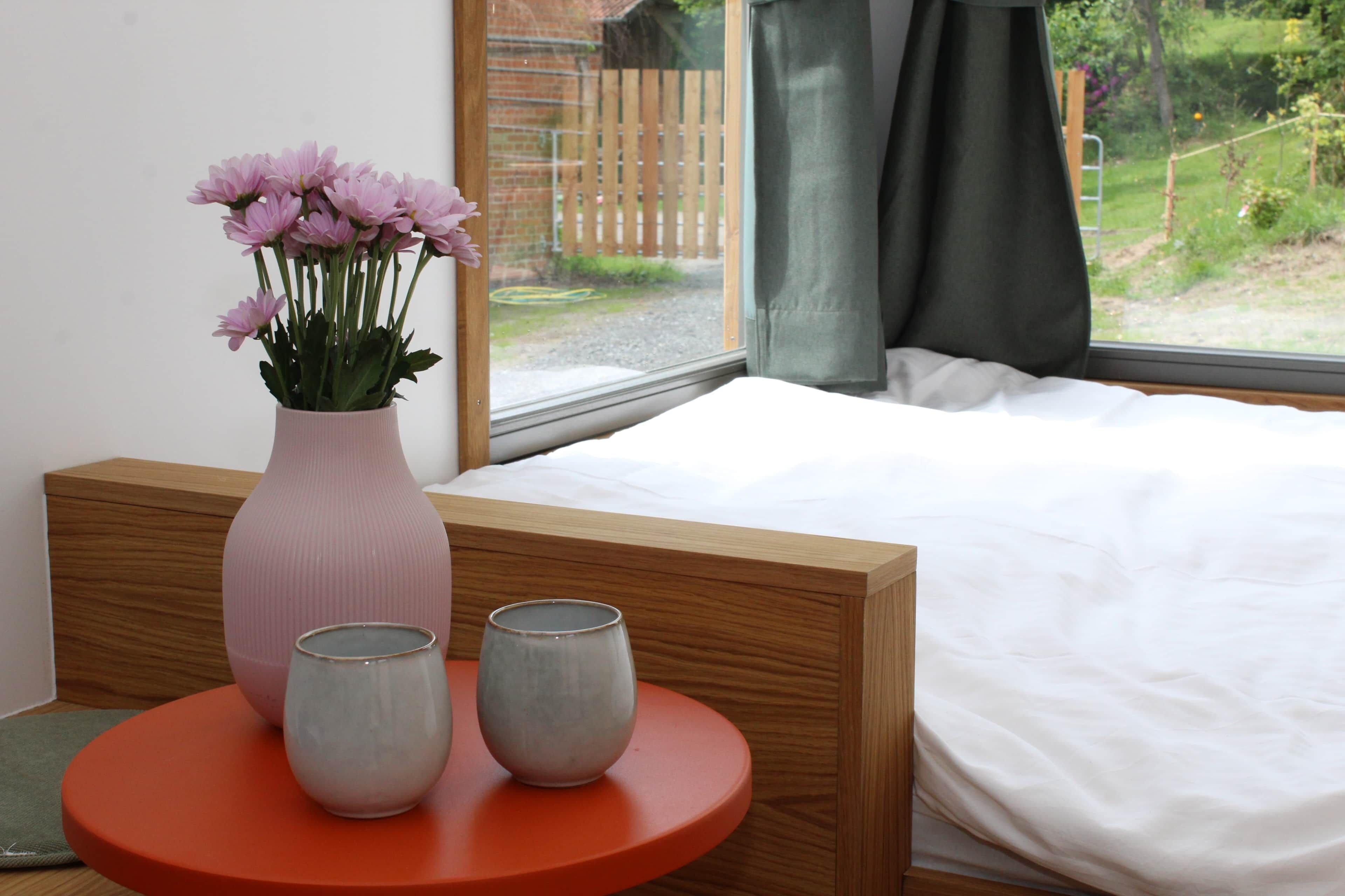 Um quarto com cama, janela com cortinas, um vaso com flores e duas xícaras de cerâmica sobre uma mesa redonda.