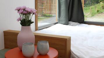 Um quarto com cama, janela com cortinas, um vaso com flores e duas xícaras de cerâmica sobre uma mesa redonda.