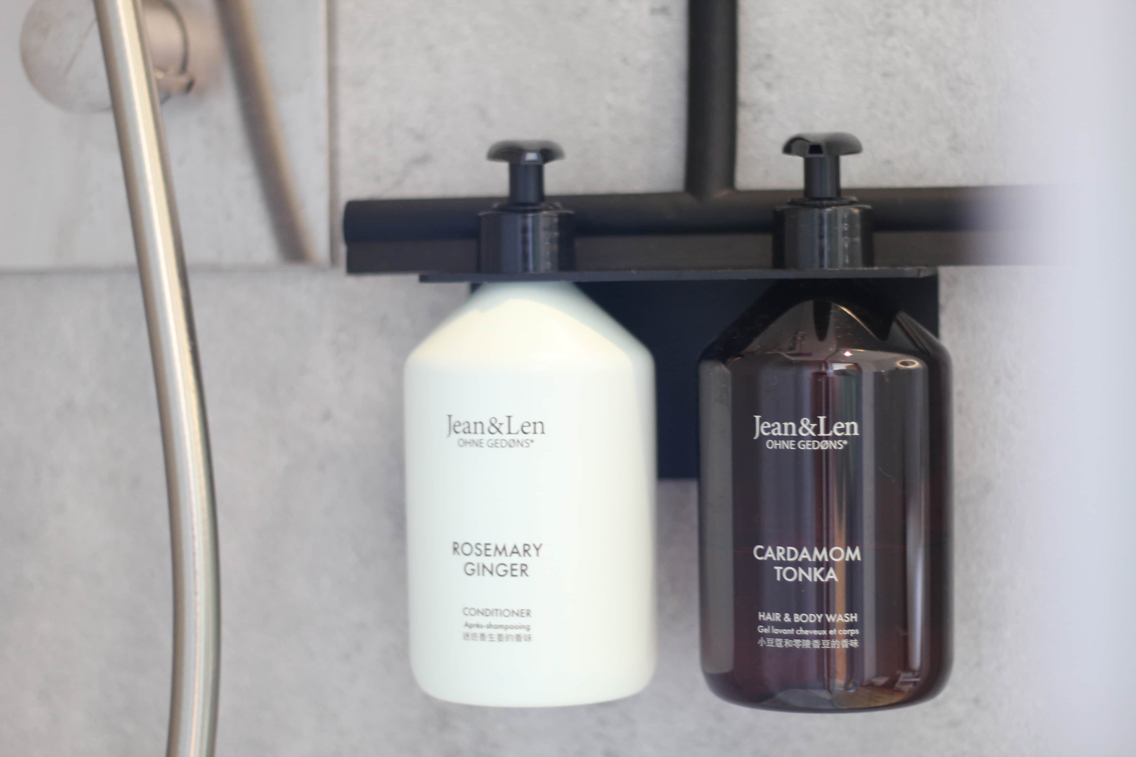 Dos botellas de productos Jean & Len, una etiquetada 'ROSEMARY GINGER' y la otra 'CARDAMOM TONKA', sobre un estante negro en un baño.