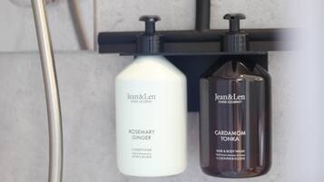 Dos botellas de productos Jean & Len, una etiquetada 'ROSEMARY GINGER' y la otra 'CARDAMOM TONKA', sobre un estante negro en un baño.
