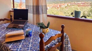 A laptop on a table with a book and a mug, a chair, and a view of a town through a window. - Beautiful private villa for 5 people with WIFI, A/C, TV, terrace and panoramic view (Villasimius)