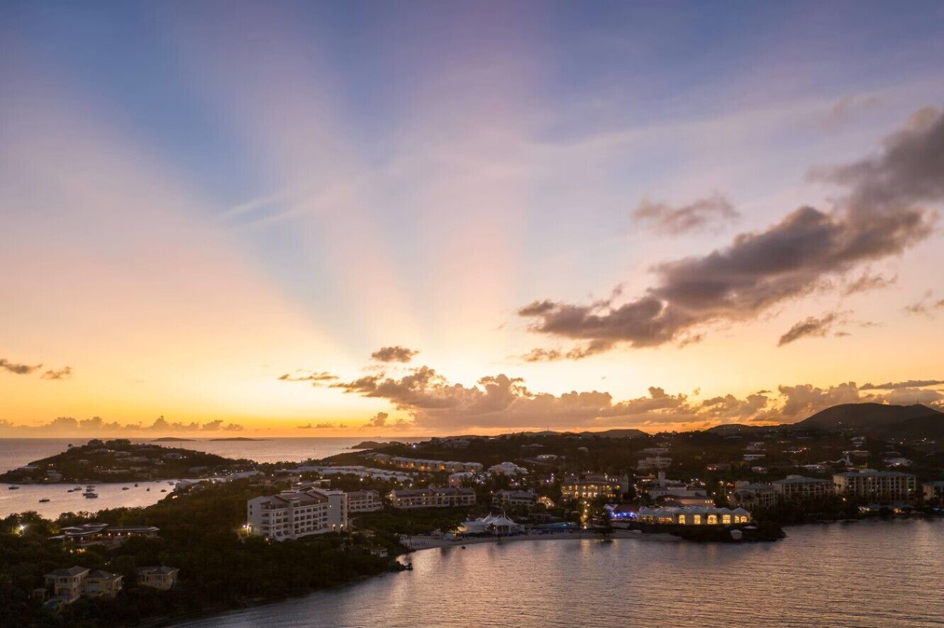 A coastal cityscape at sunset with buildings along the waterfront, a marina with boats, and a prominent hill in the background.