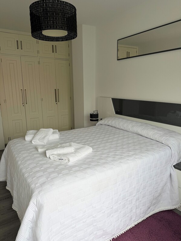 A neatly made bed with white linens and towels, a black hanging lamp, a large mirror, and a wardrobe with multiple doors.