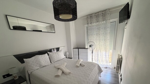 Ein Schlafzimmer mit einem Bett, einem Nachttisch, einer Lampe, einem Ventilator, einem Spiegel und einem Fenster mit Vorhängen.