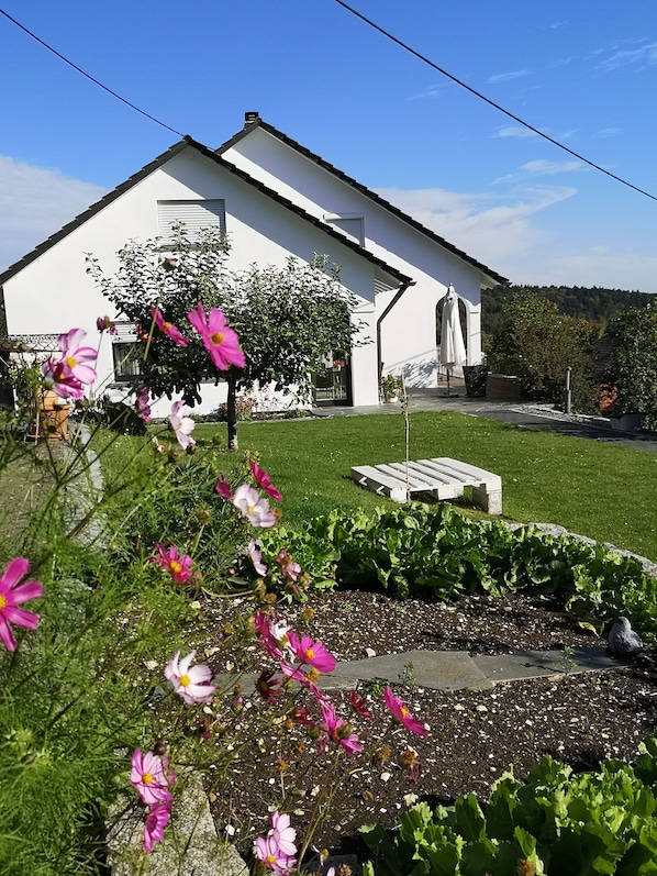 Un bâtiment blanc avec un toit en pente, un jardin parsemé de fleurs roses et un ciel d’un bleu limpide.