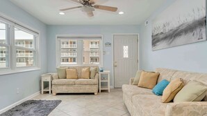 A living room with a beige sofa, a white side table, a grey rug, a ceiling fan, and a large window with a view of a building. - Oceanview Beach Retreat (Wildwood Crest)