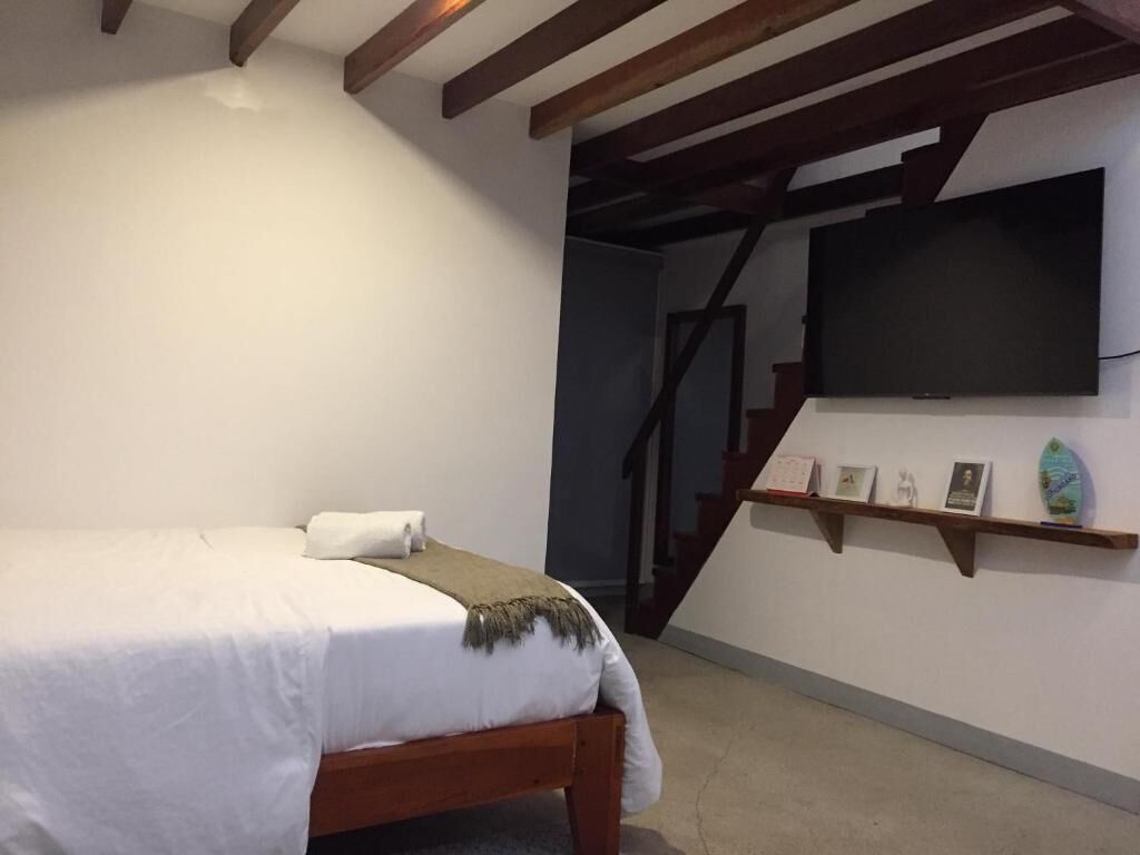 A bedroom with a bed, a TV mounted on the wall, a staircase, and a shelf with framed items.