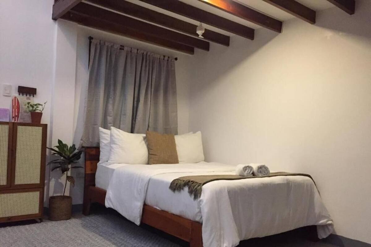 Ein Schlafzimmer mit Bett, Holz-Kopfstück, Fenster mit Vorhängen, einer kleinen Pflanze und einem an der Wand befestigten Regal.