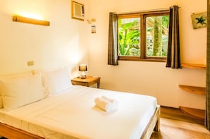 A hotel room with a bed, a nightstand, a lamp, a window with curtains, and a wooden shelf. - Amihan Siargao (General Luna)