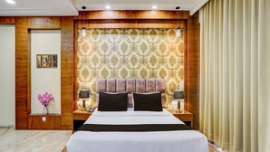 Hotellihuone, jossa on suuri sänky, puinen pääty, kuvioitu tapetti, kaksi yöpöytää valaisimineen, maljakko kukkineen ja peilillä varustettu ovi.