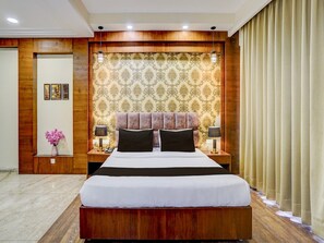 Ein Hotelzimmer mit einem großen Bett, einem Holzkopfstück, gemustertem Wandpapier, zwei Nachttischen mit Lampen, einer Blumenvase und einer Tür mit Spiegel.